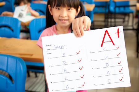 smiling little girl showing exam paper with a plus in the classroom